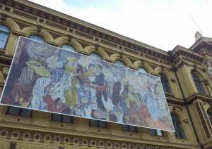 Monumentaaliteos Juhlat kaupungissa koristaa ateneumin julkisivua. Tove Jansson vetää röökiä kuvan etuosassa.